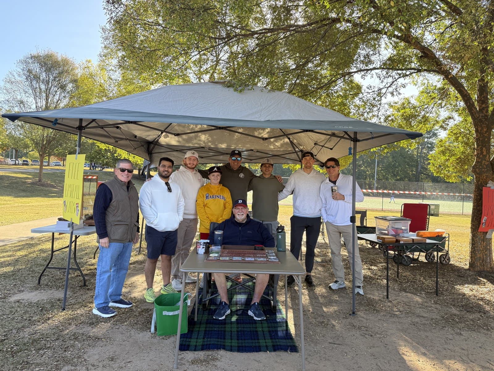 The CardFather seated at his card table under a tent, family and friends standing behind him.